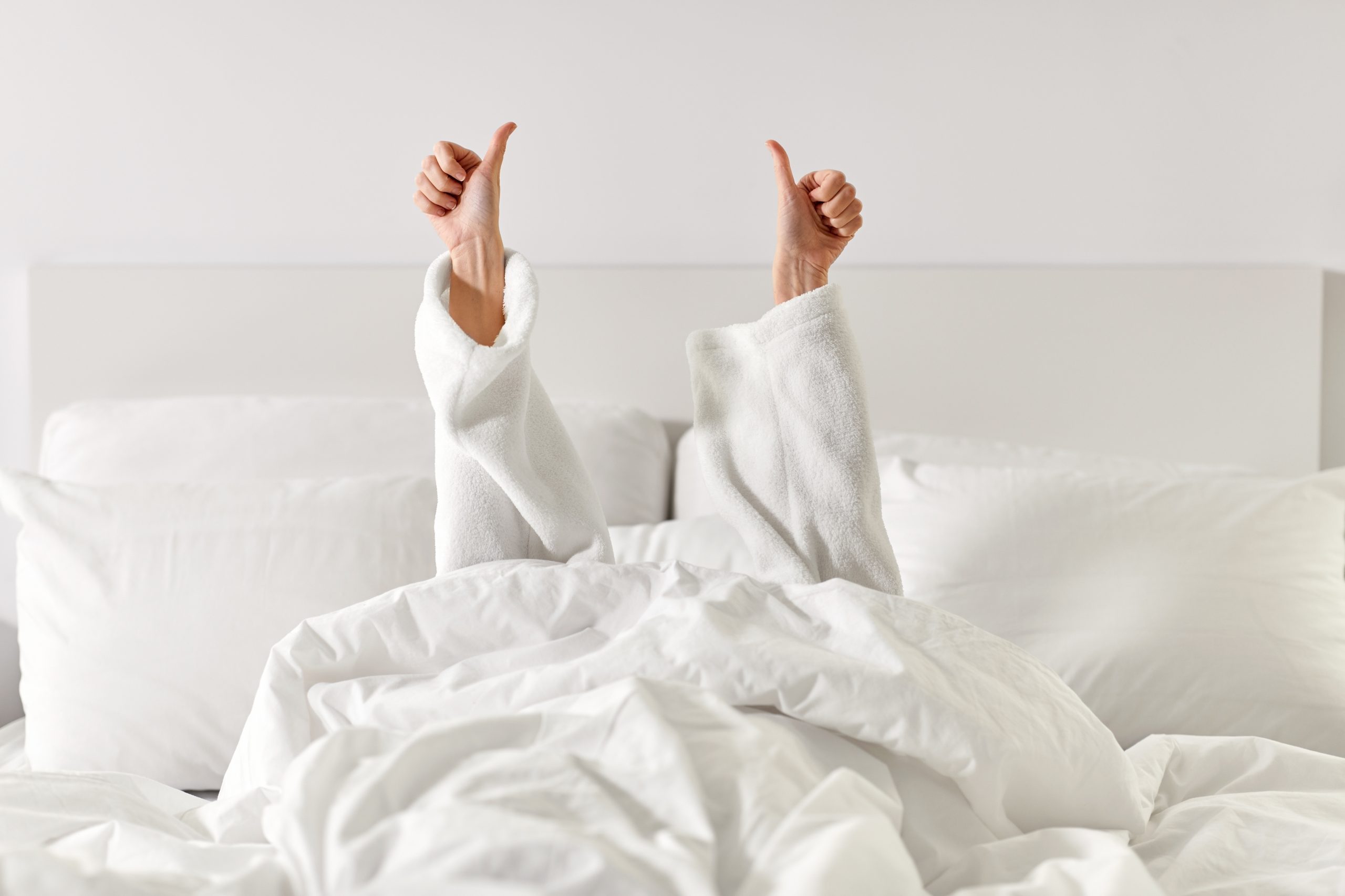 Eine Person liegt unter einer weissen Decke im Bett und streckt beide Daumen hoch, gekleidet in einen weissen Bademantel. Das Bild vermittelt ein Gefühl von Erholung und positiver Energie. Guter Schlaf kann bei ADHS Schweiz eine wichtige Rolle für die Tagesstruktur und das Wohlbefinden spielen.