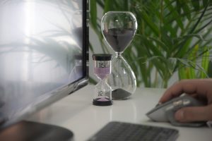 Zwei Sanduhren stehen neben einem Computerbildschirm auf einem Schreibtisch, während eine Hand zur Maus greift. Die Sanduhren symbolisieren Zeitmanagement und einen strukturierten Umgang mit Zeit.