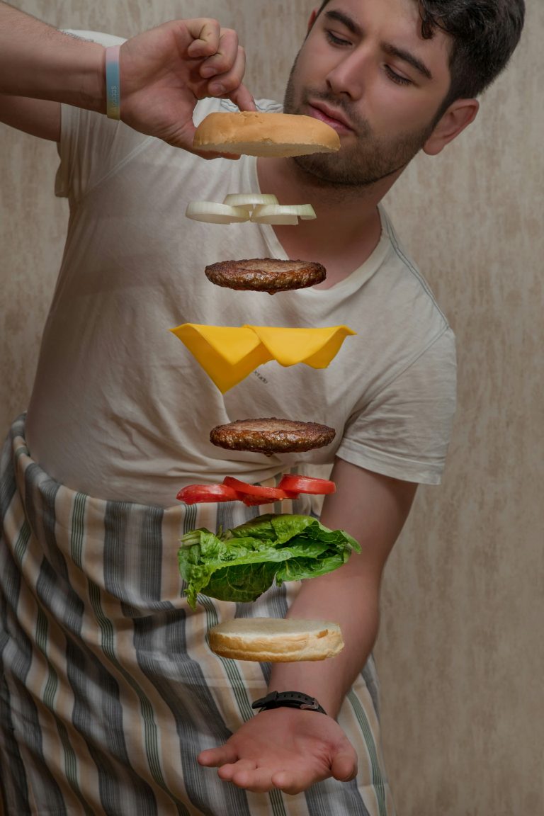 Ein Mann hält mit einer Hand mehrere Burger-Zutaten in der Luft, darunter Salat, Tomaten, Käse und Brötchen. Ernährung spielt auch bei ADHS Schweiz eine wichtige Rolle, da bestimmte Nahrungsmittel die Symptomatik positiv oder negativ beeinflussen können. Eine ausgewogene Ernährung unterstützt oft den Behandlungserfolg.