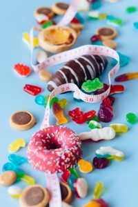 Eine bunte Auswahl an Süssigkeiten wie Donuts, Gummibärchen und Schokolade liegt auf einem hellblauen Untergrund, umwickelt von einem Massband.