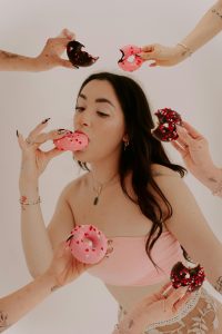 Eine junge Frau isst genussvoll einen pinken Donut, während mehrere Hände ihr weitere Donuts anbieten.
