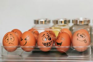 Braune Eier mit unterschiedlichen, aufgemalten Gesichtsausdrücken stehen in einer durchsichtigen Halterung. Das Bild steht sinnbildlich für die Vielfalt der Gefühle und ihr schnelles Wechseln.