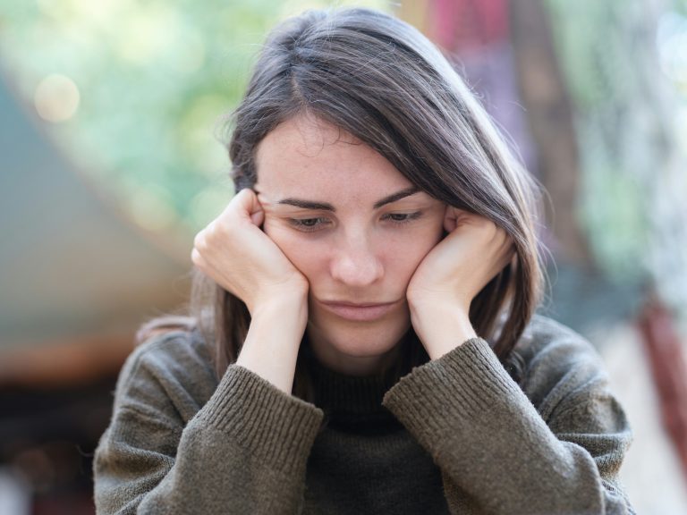 Eine junge Frau sitzt nachdenklich mit dem Kopf in den Händen. Der Ausdruck spiegelt innere Erschöpfung, emotionale Überforderung und das Gefühl, mit Gedanken festzustecken – typisch für ADHS im Alltag.