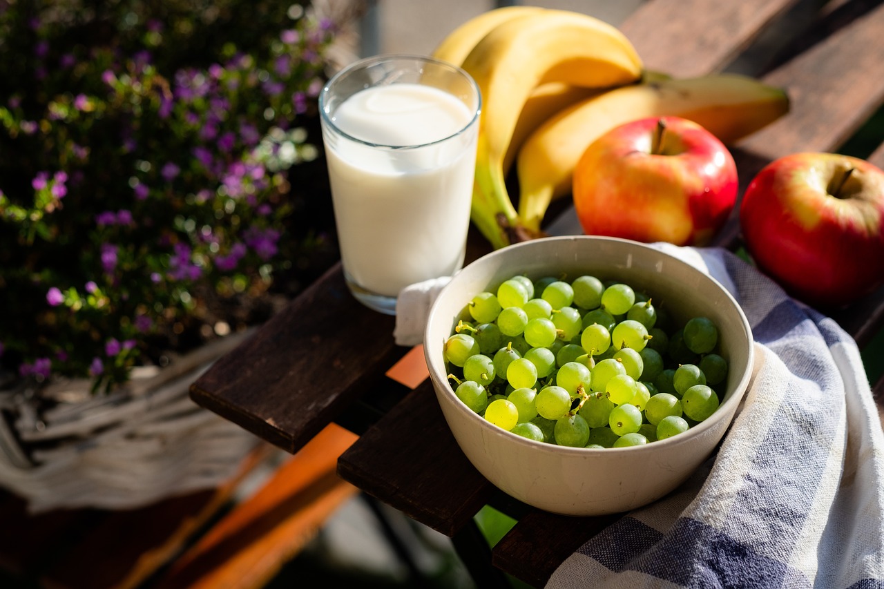 Ein Glas Milch, eine Schale voller grüner Trauben, Bananen und rote Äpfel stehen auf einem Holztisch im Freien. Frisches Obst kann einen positiven Einfluss auf die Schlafqualität bei ADHS Schweiz haben. Eine ausgewogene Ernährung unterstützt die Regulation des Energiehaushalts und der Konzentration.