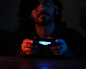 Ein junger Mann hält einen leuchtenden Controller und schaut nachdenklich ins Dunkel. Das blaue Licht wirft eine fast meditative Stimmung.