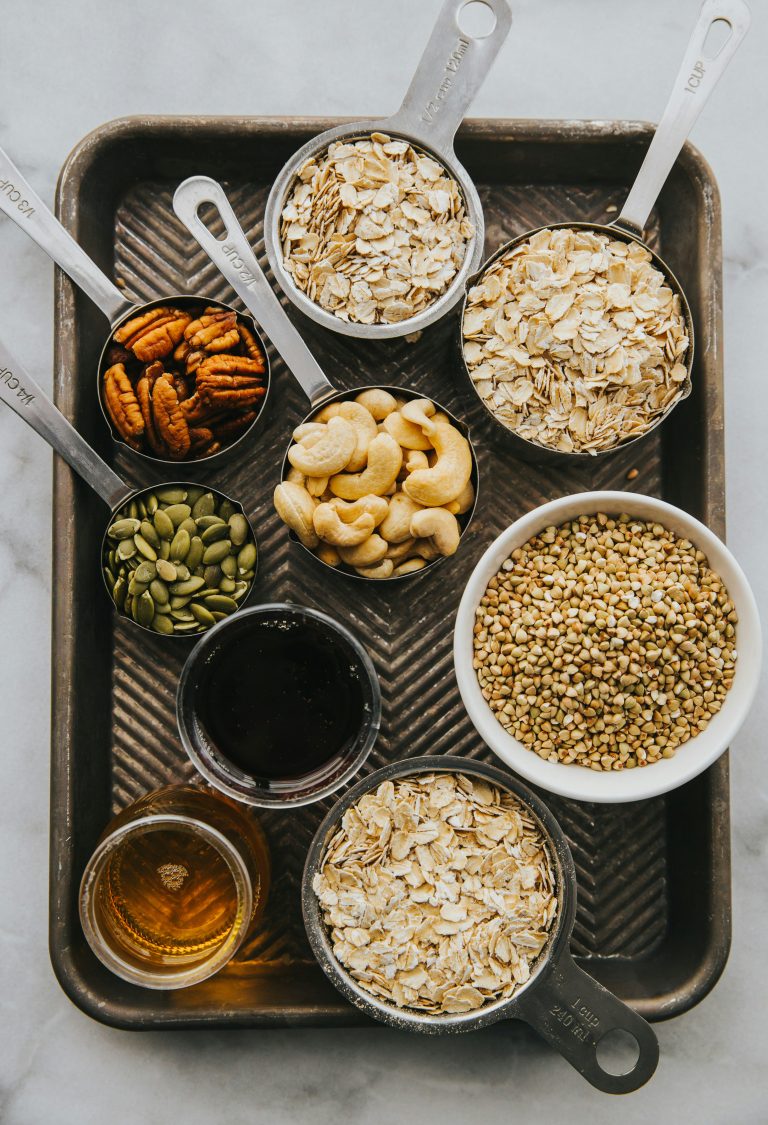 Mehrere Messbecher und Schalen sind mit Haferflocken, Cashewkernen, Kürbiskernen und Buchweizen gefüllt und liegen ordentlich auf einem Backblech. Auch kleine Gläser mit Honig und Sirup sind zu sehen. Eine ausgewogene Ernährung wie diese wird in ADHS Schweiz oft als begleitende Massnahme zur Therapie empfohlen.