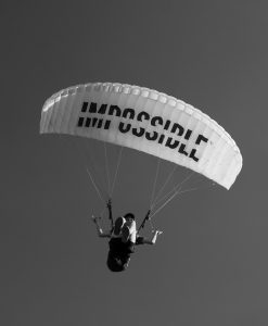 Ein Fallschirmspringer fliegt unter einem Schirm mit der Aufschrift „IMPOSSIBLE“, wobei das Wort durch die Position des Körpers als „POSSIBLE“ gelesen werden kann. Das Bild steht für das Überwinden innerer Grenzen.