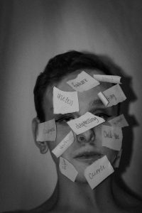 Schwarz-weiss Porträt eines Jugendlichen mit zahlreichen beschrifteten Zetteln im Gesicht, Wörter wie „failure“, „useless“ oder „depressing“ symbolisieren das Stigma, die Selbstzweifel und komorbiden Symptome wie Depression bei ADHS.