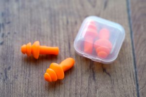 Drei orangefarbene Ohrstöpsel liegen auf einem Holztisch, daneben eine kleine Plastikbox. Ohrstöpsel können störende Geräusche im Schlafzimmer dämpfen.