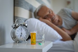 Ein älterer Mann schläft friedlich im Bett, auf dem Nachttisch stehen ein Wecker, ein Glas Wasser und eine Dose Medikamente.
