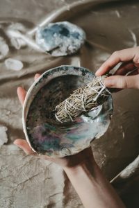 Eine Hand hält ein traditionelles Räucherbündel über einer Muschelschale, ein Ritual, das mit Naturheilkunde und Entspannung in Verbindung gebracht wird.