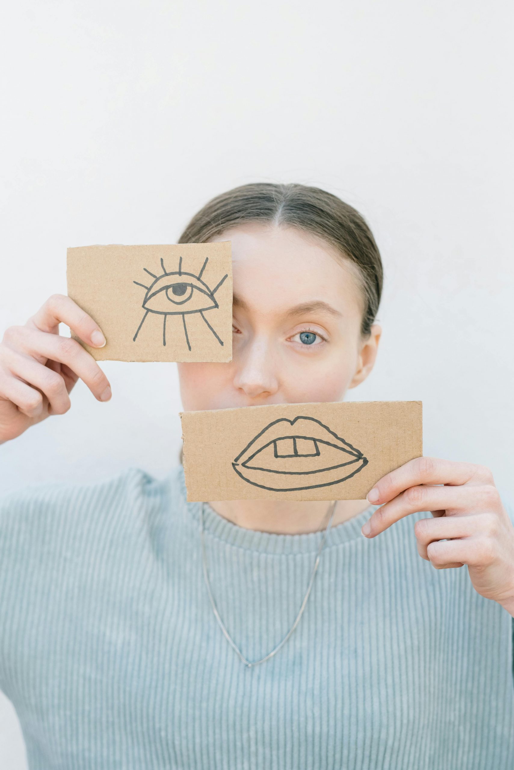 Eine junge Frau hält Pappstücke mit einem gezeichneten Auge und Mund vor ihr Gesicht – ein Symbol für die Schwierigkeit, Emotionen bei ADHS oder Autismus auszudrücken.