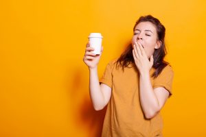 Eine junge Frau gähnt müde und hält einen Kaffeebecher in der Hand vor einem knallgelben Hintergrund. Müdigkeit ist ein häufiges Problem bei ADHS, da Schlafprobleme zu Konzentrationsschwierigkeiten führen können. Koffein ersetzt keinen gesunden Schlaf.
