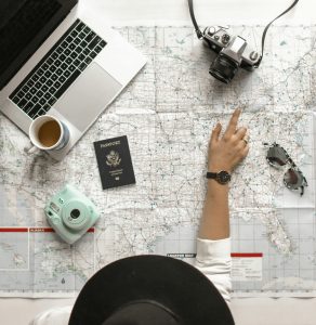 Eine Person plant ihre nächste Reise und zeigt auf eine Landkarte, umgeben von Laptop, Kamera, Pass und Kaffee.