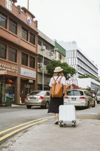 Eine Frau mit Hut und Rucksack zieht einen Koffer durch eine geschäftige Stadtstrasse.