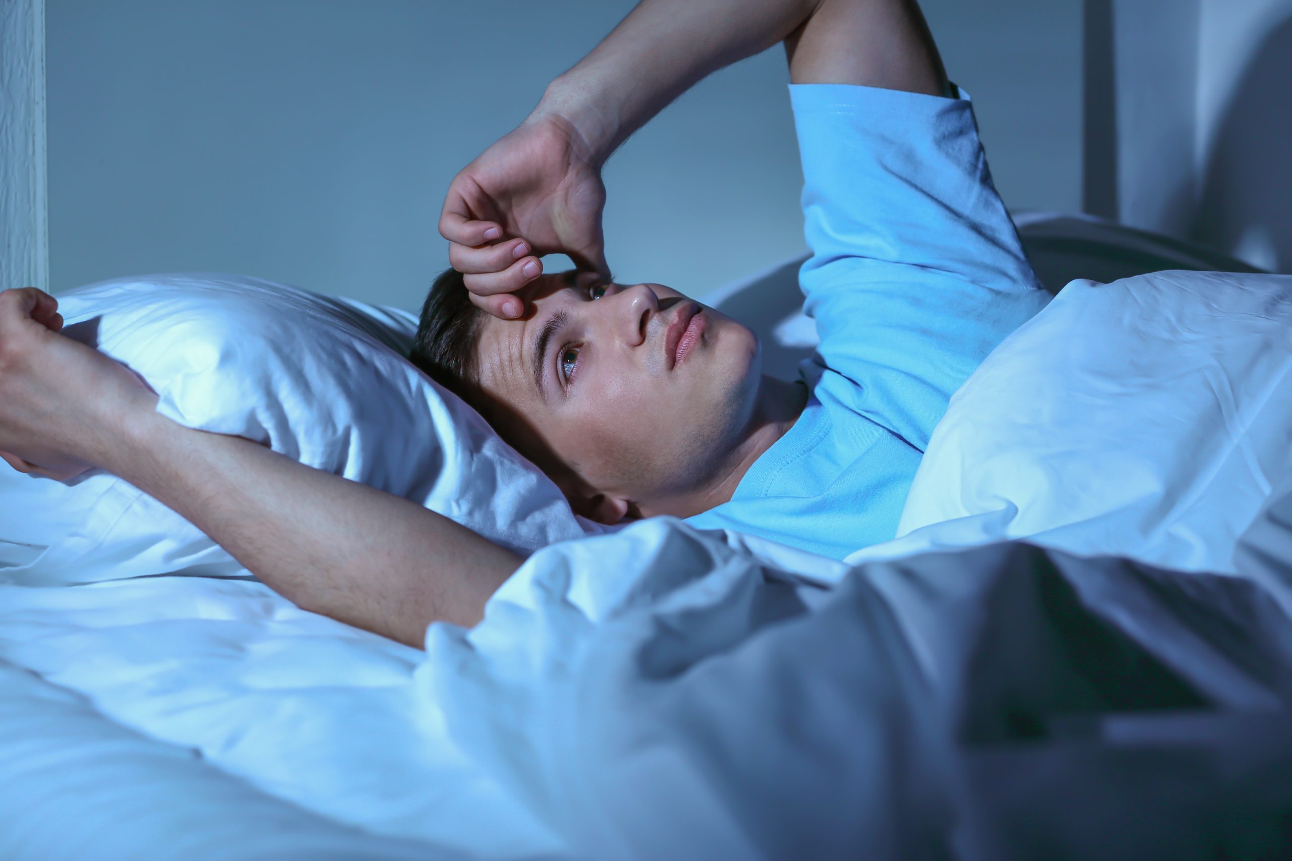 Ein junger Mann liegt nachts wach im Bett, schaut gedankenverloren an die Decke und wirkt angespannt. Das kühle Licht des Zimmers betont seine Schlaflosigkeit. Einschlafprobleme gehören bei ADHS Schweiz zu den typischen Begleiterscheinungen.
