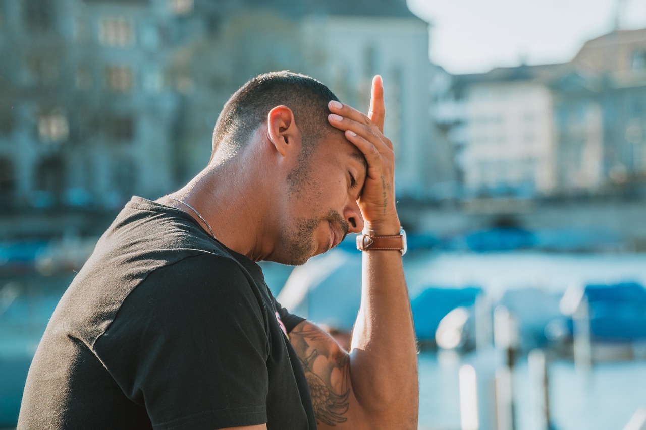 Ein Mann sitzt im Freien und hält sich frustriert die Stirn, im Hintergrund ist eine Stadt mit Wasser und Booten zu sehen. Auch bei ADHS Schweiz ist emotionaler Stress ein zentrales Thema, das sich körperlich und seelisch zeigen kann. Pausen und Selbstfürsorge sind wichtige Elemente der Therapie.