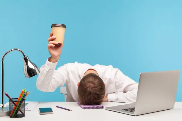 Ein Mann in weissem Hemd liegt erschöpft auf seinem Schreibtisch, während er einen Pappbecher in die Höhe hält. Neben ihm stehen ein Laptop, ein Handy und Büromaterialien. Müdigkeit und Konzentrationsprobleme sind typische Herausforderungen für Menschen mit ADHS Schweiz.